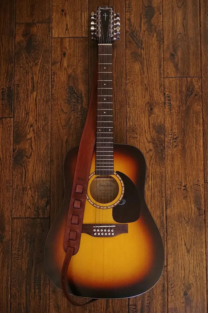 A acoustic guitar with a leather camera strap laying on a wooden floor.