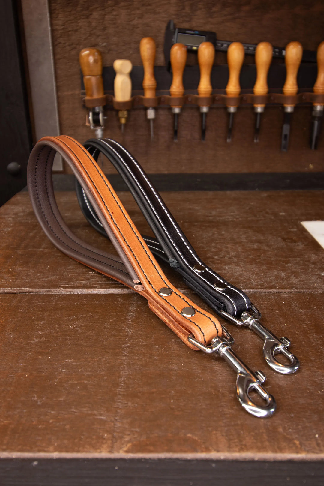 Two leather dog leashes on a wooden surface with tools in the background.