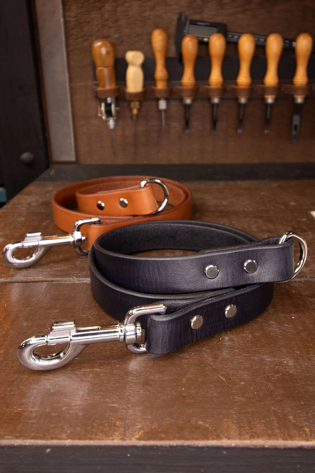 Two leather leash lengths, one black and one brown, on a wooden surface with tools in the background.