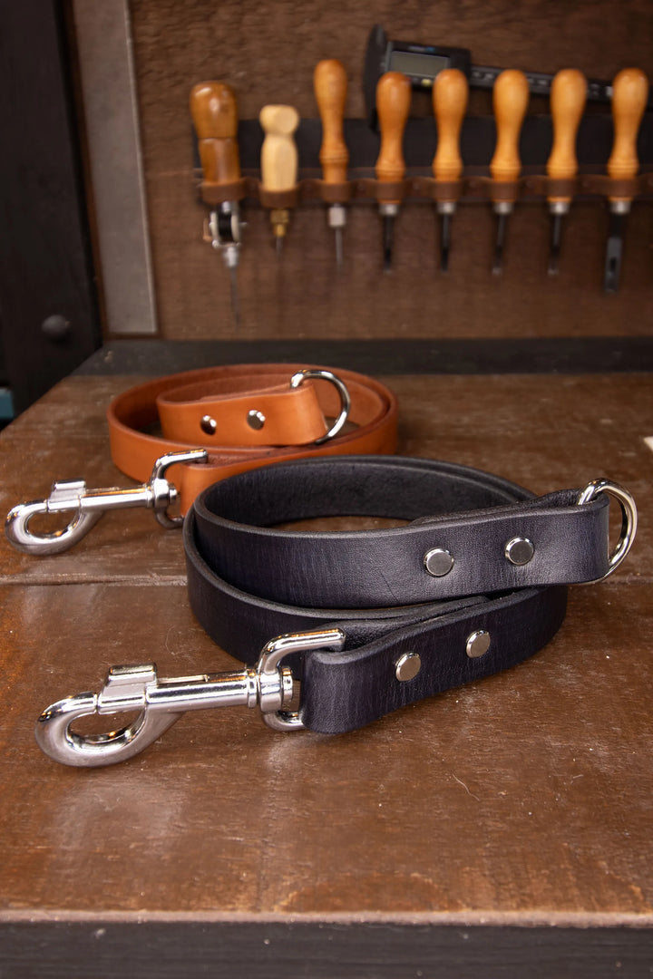 Two leather leash lengths, one black and one brown, on a wooden surface with tools in the background.