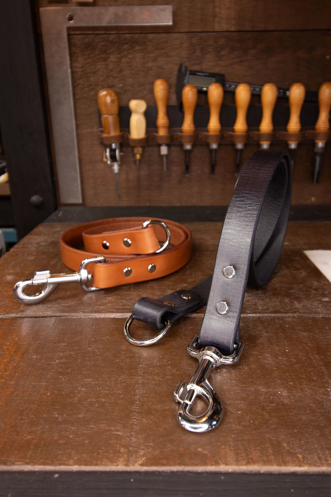 Two leather leash lengths ad-on with metal clasps and metal D-rings on a wooden surface with tools in the background.