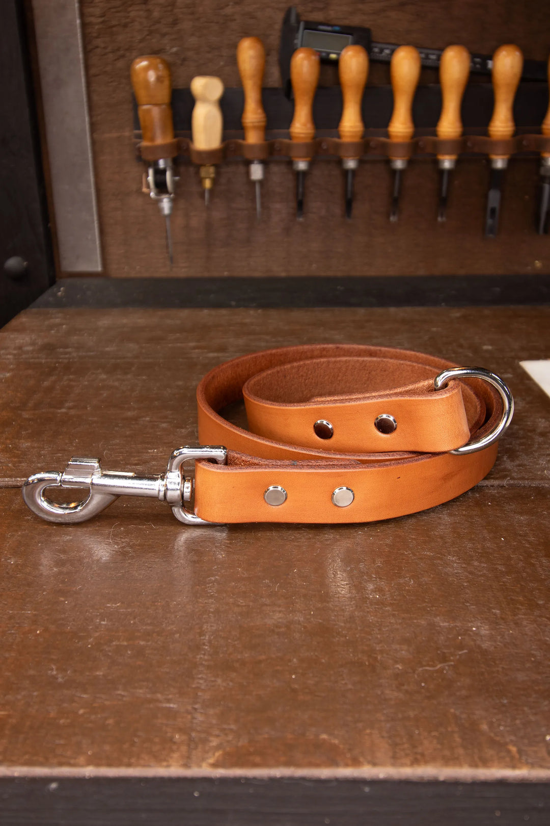 Brown leather dog leash with silver clip on one end and a D-ring on the other end on a wooden surface with tools in the background