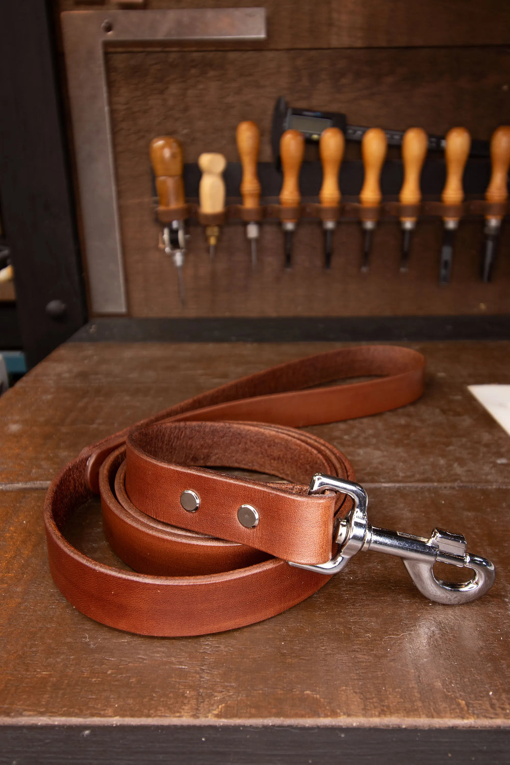 Brown leather dog leash with metal clip on a wooden surface with tools in the background