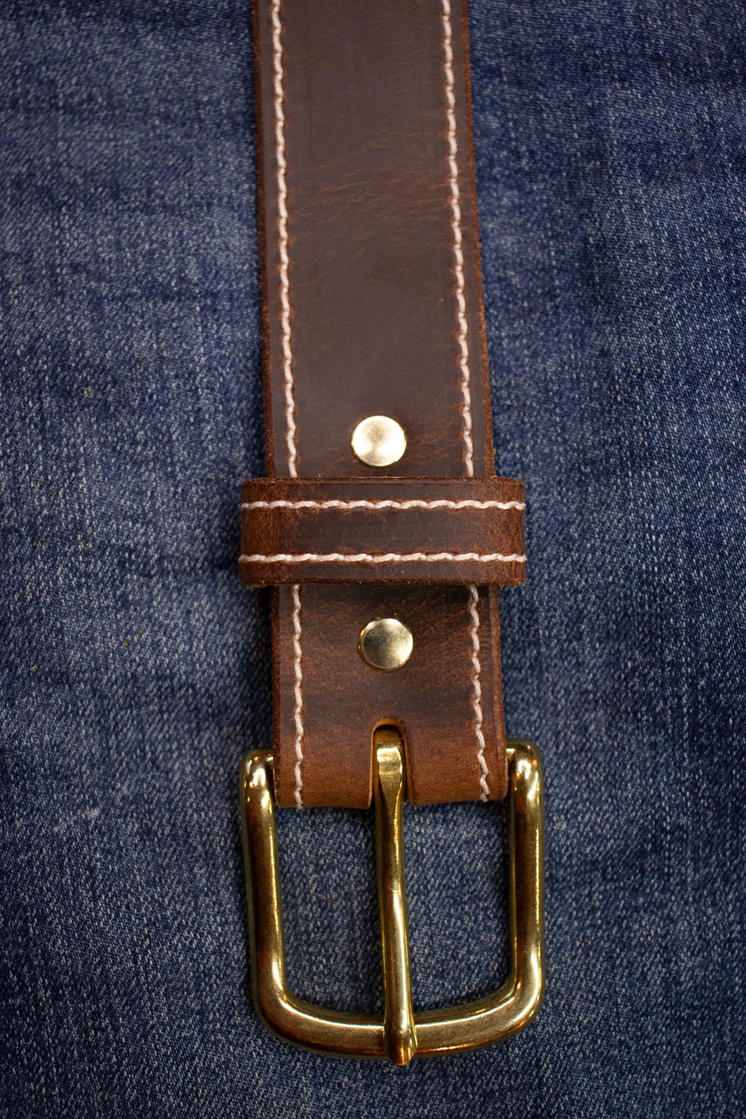 Brown leather belt with thick stitching with brass buckle on a denim background